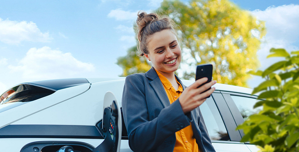 Frau mit Smartphone in der Hand lehnt an ihr Elektroauto, das gerade geladen wird