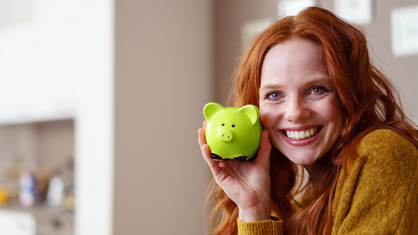 Eine rothaarige Frau lächelt und hält ein grünes Sparschwein hoch.
